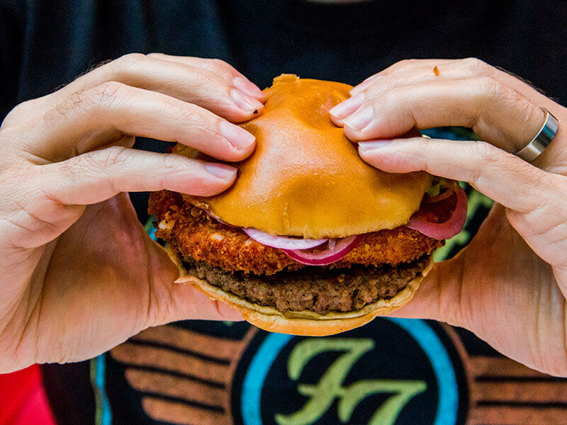 Burger da casa nas mãos, com carne, cebola roxa e queijo empanado